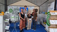 Eine Gruppe von vier Personen steht im Vordergrund eines Messestands. Im Hintergrund sind Informationsmaterialien und Banner zu sehen. Die Personen lächeln und tragen Namensschilder. Die Szenerie vermittelt eine freundliche und einladende Atmosphäre.