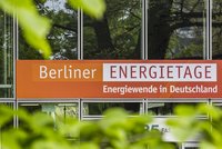 Ein bannerförmiger Schriftzug zeigt die Wörter "Berliner ENERGIEtage" auf orangefarbenem Hintergrund, darunter steht "Energiewende in Deutschland". Der Schriftzug ist vor einer modernen Glasfassade platziert, umgeben von grünen Blättern.