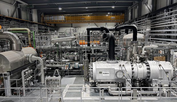 Das Bild zeigt eine industrielle Anlage mit zahlreichen Rohren, Maschinen und Tanks. Im Vordergrund befindet sich eine große Maschine mit mehreren Rohren, während im Hintergrund weitere Anlagen und eine steinerne Plattform zu sehen sind. Die Umgebung ist gut beleuchtet und wirkt modern.