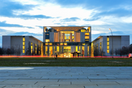 Das Bild zeigt ein modernes Gebäude mit einer beeindruckenden Architektur, beleuchtet in den Abendstunden. Die Fassade besteht aus einem Mix aus Beton und Glas. Im Vordergrund ist ein Platz mit Pflastersteinen und einige grüne Flächen zu sehen.