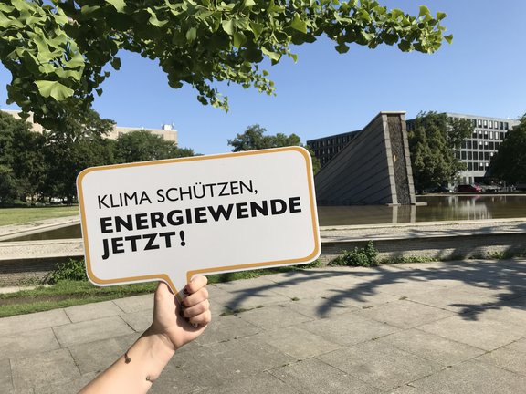 Eine Hand hält ein großes Schild mit der Aufschrift: "KLIMA SCHÜTZEN, ENERGIEWENDE JETZT!" Die Szene spielt sich in einem Park mit Bäumen und Wasser im Hintergrund ab.