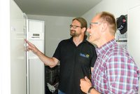 Zwei Männer stehen vor einem Kühlschrank. Der Mann links zeigt mit dem Finger auf ein Bedienelement, während der Mann rechts freundlich lächelt. Beide scheinen ein Gespräch über den Kühlschrank zu führen.