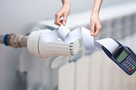 Eine Hand hält ein Stück Papier, das von einem Heizkörper herab hängt, während die andere Hand ein digitales Thermostat zeigt. Die Darstellung vermittelt einen Bezug zur Heizungssteuerung und Ablesung von Heizdaten.