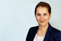 Eine junge Frau mit blonde, fleißig geflochtenem Haar lächelt freundlich in die Kamera. Sie trägt ein dunkles Blazer und eine helle Bluse. Der Hintergrund ist hell und neutral. Der Fokus liegt auf ihrem Gesicht und Ausdruck.