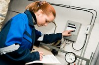 Eine Person in blauer Arbeitskleidung steht vor einem Gerätepanel an der Wand und drückt einen Knopf. In der linken Hand hält sie ein Notizbuch. Im Hintergrund sind Kabel und ein Manometer zu sehen.