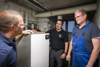 Drei Männer stehen in einem technischen Raum und unterhalten sich. Einer von ihnen zeigt auf ein Gerät, das offensichtlich eine moderne Heizungsanlage ist. Alle drei tragen Arbeitskleidung und wirken konzentriert und freundlich während des Gesprächs über die Technik.