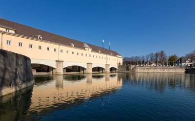 Ein historisches Gebäude mit mehreren Bogenfenstern spiegelt sich im ruhigen Wasser eines Kanals wider. Der klare, blaue Himmel und die umliegenden Bäume sind im Hintergrund sichtbar.