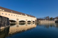 Ein historisches Gebäude mit mehreren Bogenfenstern spiegelt sich im ruhigen Wasser eines Kanals wider. Der klare, blaue Himmel und die umliegenden Bäume sind im Hintergrund sichtbar.