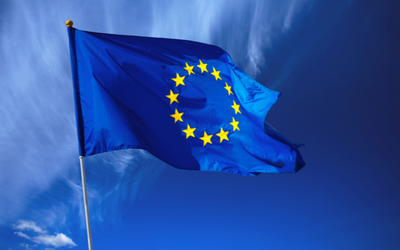 Eine blaue Flagge mit einem Kreis aus zwölf gelben Sternen in der Mitte, die die Europäische Union symbolisiert. Der Hintergrund zeigt einen klaren blauen Himmel mit einigen leichten Wolken.