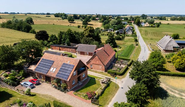 Luftaufnahme eines ländlichen Wohngebiets mit mehreren Häusern. Im Vordergrund steht ein Haus mit Solarpanelen auf dem Dach, umgeben von einem gepflegten Garten. Eine ruhige Straße verläuft entlang der Nachbarhäuser und Wiesen, die die Landschaft dominieren.