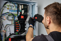 Ein Techniker arbeitet an einem Schaltschrank. Er trägt schwarze Handschuhe und benutzt einen Schraubendreher, um Kabel und Komponenten in einem elektrischen Verteiler zu überprüfen oder zu reparieren. Der Bereich ist mit verschiedenen Drähten und Geräten voll sichtbar.