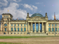 Das Bild zeigt die Fassade des Reichstagsgebäudes in Berlin. Es ist ein historisches, monumental gestaltetes Gebäude mit einer großen Treppe und einer Glaskuppel. Über dem Haupteingang steht „Dem Deutschen Volke“. Im Hintergrund sind einige Flaggen zu sehen.