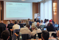 Ein Vortragender steht vor einem Publikum in einem Konferenzraum. Im Hintergrund ist eine Präsentation zu sehen, die Informationen zu HEMS und Wärmequellen zeigt. Die Zuhörer sind sitzend und auf den Vortrag konzentriert.