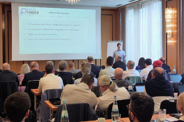 Ein Vortragender steht vor einem Publikum in einem Konferenzraum. Im Hintergrund ist eine Präsentation zu sehen, die Informationen zu HEMS und Wärmequellen zeigt. Die Zuhörer sind sitzend und auf den Vortrag konzentriert.