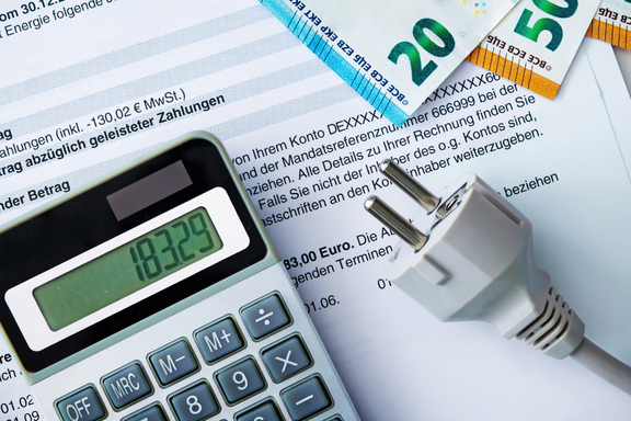 Ein Taschenrechner zeigt den Wert 183,29 an. Daneben liegt ein Stromstecker, und im Hintergrund sind Geldscheine (20 und 50 Euro) sowie ein Dokument mit Informationen zu Zahlungen zu sehen.