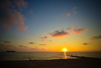Sonnenuntergang über dem Meer, mit orangefarbenem Himmel und einigen Wolken. Am Horizont sind mehrere Silhouetten von Menschen zu sehen, die auf einem Steg stehen. Im Hintergrund ist ein Schiff auf dem Wasser sichtbar.