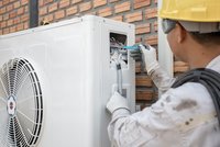 Ein Techniker in Arbeitskleidung und Handschuhen überprüft eine Klimaanlage. Er verwendet einen blauen Werkzeuggriff, um Kabel im Inneren des Geräts zu verbinden. Im Hintergrund ist eine Ziegelwand sichtbar.