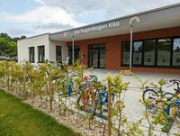 Eine Kita namens "Die Regenbogen Kita" mit einem modernen Eingangsbereich. Vor dem Gebäude stehen bunt lackierte Kinderräder und ein grüner Heckenzaun. Die Fenster zeigen Zeichnungen von Kinderfiguren.