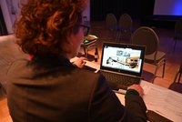 Eine Person mit lockigem Haar sitzt an einem Tisch und schaut auf einen Laptop. Auf dem Bildschirm sind mehrere Teilnehmer einer Videokonferenz sichtbar. Auf dem Tisch liegen Dokumente, und im Hintergrund sind leere Stühle zu sehen.