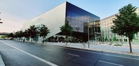 Das Bild zeigt das Futurium, ein modernes, architektonisch gestaltetes Gebäude mit einer glatten, schimmernden Fassade. Vor dem Gebäude befinden sich Bäume und ein Gehweg. Im Vordergrund verläuft eine Straße mit einem Fahrrad und einem Straßenlaterne.