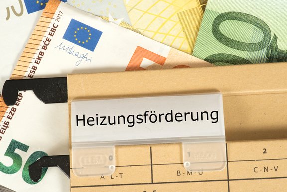Ein Ordner mit dem beschrifteten Tab „Heizungsförderung“ liegt auf einem Untergrund aus verschiedenen Euro-Banknoten. Die Banknoten sind teilweise sichtbar und verleihen der Szene einen finanziellen Kontext.