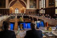 Eine große Konferenz mit zahlreichen Teilnehmern in einem historischen Saal. An einem langen Tisch befinden sich Bildschirme, auf denen ein Redner zu sehen ist. Blumenarrangements und Getränke sind auf dem Tisch verteilt, während viele Personen aufmerksam zuhören und Notizen machen.