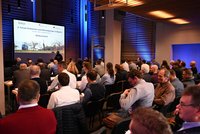 Eine Gruppe von Fachleuten sitzt in einem Konferenzraum, während auf einer großen Leinwand die Begrüßung für das 2. Forum zu Erdwärme und Wärmepumpen in Bayern angezeigt wird. Der Raum ist modern gestaltet und in der ersten Reihe steht ein Redner.