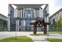 Das Bild zeigt das Bundeskanzleramt in Berlin, ein modernes Gebäude mit einer markanten Architektur aus Glas und Beton. Im Vordergrund steht eine rustikale Skulptur. Der Bereich ist mit einem Zaun umgeben und es gibt gepflegte Rasenflächen.