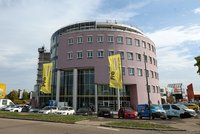 Ein modernes, kreisförmiges, rosa Gebäude mit großen Fenstern und einer Glasfront. Vor dem Gebäude sind verschiedene PKWs geparkt. An der Fassade weht ein gelbes Banner mit der Aufschrift "ProMaus Unternehmensgruppe". Der Himmel ist teilweise bewölkt.