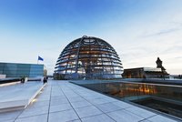 Auf diesem Bild ist die gläserne Kuppel des Deutschen Bundestages in Berlin zu sehen, umgeben von modernen Gebäuden und einem klaren blauen Himmel. Die Struktur ist futuristisch und symbolisiert Transparenz in der Politik.