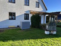 Ein einseitiges Haus mit weißer Fassade, dunklen Fenstern und einem überdachten Eingang. Im Vordergrund steht eine graue Klimaanlage neben einem kleinen, grünen Busch und einem Steinkamin. Der Rasen ist grün und gepflegt.