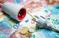 Ein Stromstecker und eine Steckdose liegen auf einem Stapel von Euro-Scheinen und einigen Münzen. Die aufliegenden Geldscheine sind in verschiedenen Farben und Werten sichtbar. Die Szene thematisiert das Zusammenspiel von Energie und Kosten.