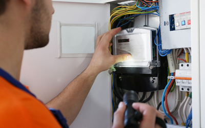 Ein Techniker mit einem Taschenlampen in der Hand überprüft einen Stromzähler in einem Elektrikerschrank, umgeben von bunten Kabeln. Die Umgebung zeigt verschiedene elektrische Komponenten und Schalter.