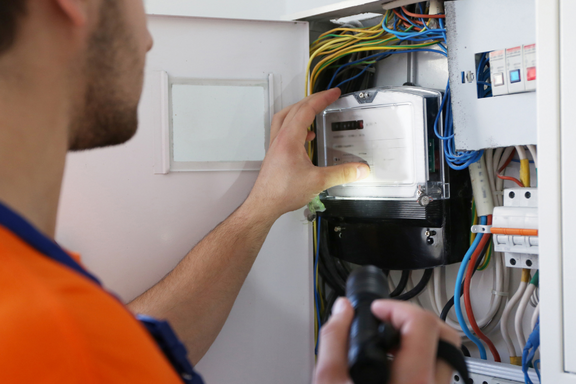 Ein Techniker mit einem Taschenlampen in der Hand überprüft einen Stromzähler in einem Elektrikerschrank, umgeben von bunten Kabeln. Die Umgebung zeigt verschiedene elektrische Komponenten und Schalter.