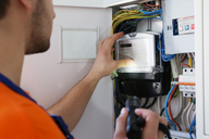 Ein Techniker mit einem Taschenlampen in der Hand überprüft einen Stromzähler in einem Elektrikerschrank, umgeben von bunten Kabeln. Die Umgebung zeigt verschiedene elektrische Komponenten und Schalter.