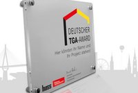 Eine transparente Tafel mit der Aufschrift „Deutscher TGA-Award“ und dem Hinweis „Hier könnten Ihr Name und Ihr Projekt stehen!“ darunter. Das Design ist modern und beinhaltet Elemente in den Farben Schwarz, Rot und Gelb. 
