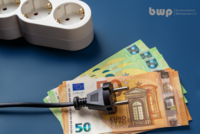 Ein Steckdosenleiste liegt auf einem blauen Tisch, daneben liegen Geldscheine in Euro, darunter ein 50-Euro-Schein. Ein Stromkabel mit Stecker ist auf die Geldscheine gelegt, was einen Zusammenhang zwischen Stromversorgung und finanziellen Aspekten suggeriert.