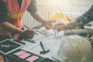 Zwei Personen in Warnwesten arbeiten gemeinsam an einem Projekt. Sie halten einen Zirkel und eine Skizze vor sich, die Baupläne zeigt. Auf dem Tisch liegen weitere Werkzeuge und Notizmaterialien. Der Fokus liegt auf der Zusammenarbeit und Planung im technischen Bereich.