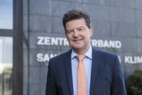 Ein Mann mit dunklen Haaren trägt einen Anzug und eine orange Krawatte. Er steht lächelnd vor einem Gebäude mit dem Schriftzug "Zentralverband Sanitär Heizung Klima". Die Atmosphäre wirkt freundlich und professionell.