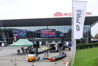 Das Bild zeigt den Eingangsbereich eines Messegeländes mit einem großen Banner, das "Willkommen" und "Eingang Nord" anzeigt. Vor dem Eingang steht ein Gabelstapler und es sind Schläuche ausgelegt. Ein Zelt ist ebenfalls zu sehen, sowie Fahnen mit dem Logo von GP JOULE.