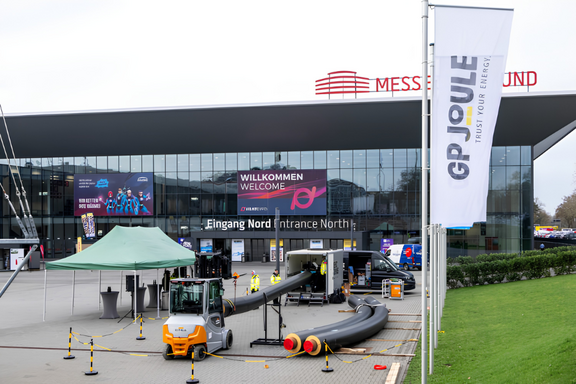 Das Bild zeigt den Eingangsbereich eines Messegeländes mit einem großen Banner, das "Willkommen" und "Eingang Nord" anzeigt. Vor dem Eingang steht ein Gabelstapler und es sind Schläuche ausgelegt. Ein Zelt ist ebenfalls zu sehen, sowie Fahnen mit dem Logo von GP JOULE.
