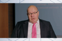 Ein Mann mit Glatze trägt eine Brille und einen dunklen Anzug mit einem pinken Hemd und Krawatte. Er spricht in die Kamera und vermittelt Informationen. Der Hintergrund ist unscharf und zeigt eine Präsentationsfläche.
