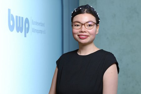 Eine lächelnde Frau mit Brille trägt ein schwarzes Kleid. Im Hintergrund ist ein Logo mit den Buchstaben "bwp" und der Aufschrift "Bundesverband Wärmepumpe e.V." zu sehen. Die Szene vermittelt ein professionelles Ambiente.