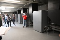 In einem modernen Raum stehen mehrere graue Geräte. Eine Person in einem roten T-Shirt und einer Maske erklärt etwas, während eine Gruppe von Menschen, einige mit Masken, aufmerksam zuhören. Eine Kamera ist auf die Person gerichtet.