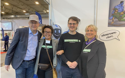 Vier Personen stehen gemeinsam vor einem Messebanner. Alle tragen T-Shirts mit der Aufschrift „Heat Pump Hero“. Im Hintergrund ist eine Cartoonfigur zu sehen, die humorvoll eine Klimaanlage präsentiert. Eine Sprechblase sagt: „Wärmepumpe: Läuft!“