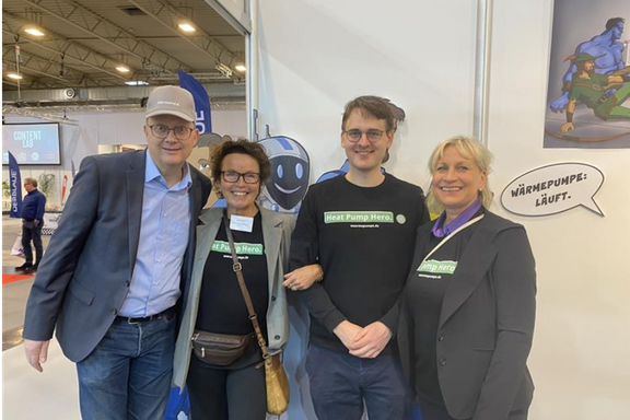 Vier Personen stehen gemeinsam vor einem Messebanner. Alle tragen T-Shirts mit der Aufschrift „Heat Pump Hero“. Im Hintergrund ist eine Cartoonfigur zu sehen, die humorvoll eine Klimaanlage präsentiert. Eine Sprechblase sagt: „Wärmepumpe: Läuft!“