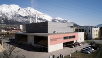 Ein modernes Biomasseheizkraftwerk in Hall in Tirol. Das Gebäude hat einen grauen oberen Teil und eine pinkfarbene Fassade. Im Hintergrund sind beeindruckende, schneebedeckte Berge zu sehen, und im Vordergrund parken mehrere Autos auf einem großen Parkplatz.