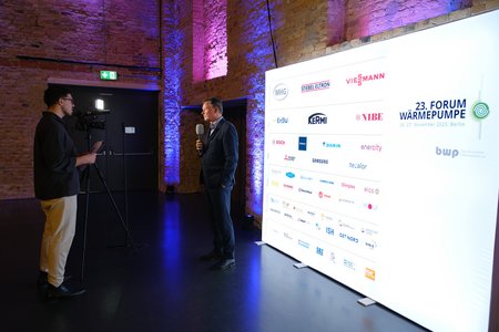 Ein Mann steht mit einem Mikrofon vor einer beleuchteten Wand, die das Logo des 23. Forums Wärmepumpe zeigt. Neben ihm steht eine Person mit einer Kamera. Der Raum ist schwach beleuchtet, mit bunten Lichtern auf den Wänden.