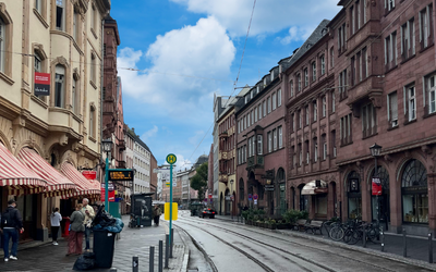 Eine lebendige Stadtstraße mit historischen Gebäuden beiderseits. Auf der linken Seite befinden sich Cafés mit Markisen. Im Vordergrund schlängelt sich eine Straßenbahnlinie. Menschen schlendern auf dem Gehweg, während Fahrräder am Straßenrand abgestellt sind. Der Himmel ist leicht bewölkt.