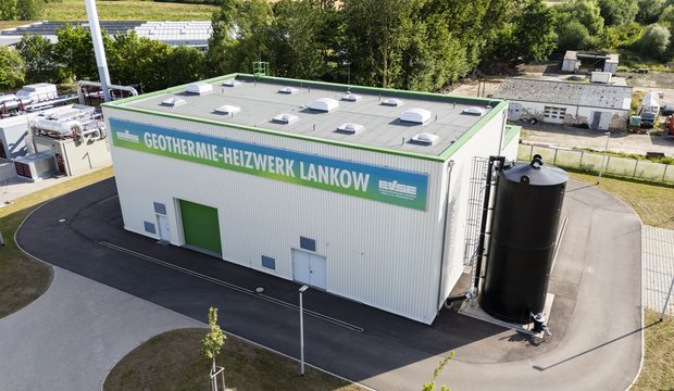 Das Bild zeigt ein Geothermie-Heizwerk in Lankow. Das Gebäude hat eine helle Fassade mit einem grünen Eingang und einem großen Schriftzug. Neben dem Gebäude steht ein schwarzer Wasserspeicher. Im Hintergrund sind Bäume und Felder sichtbar.